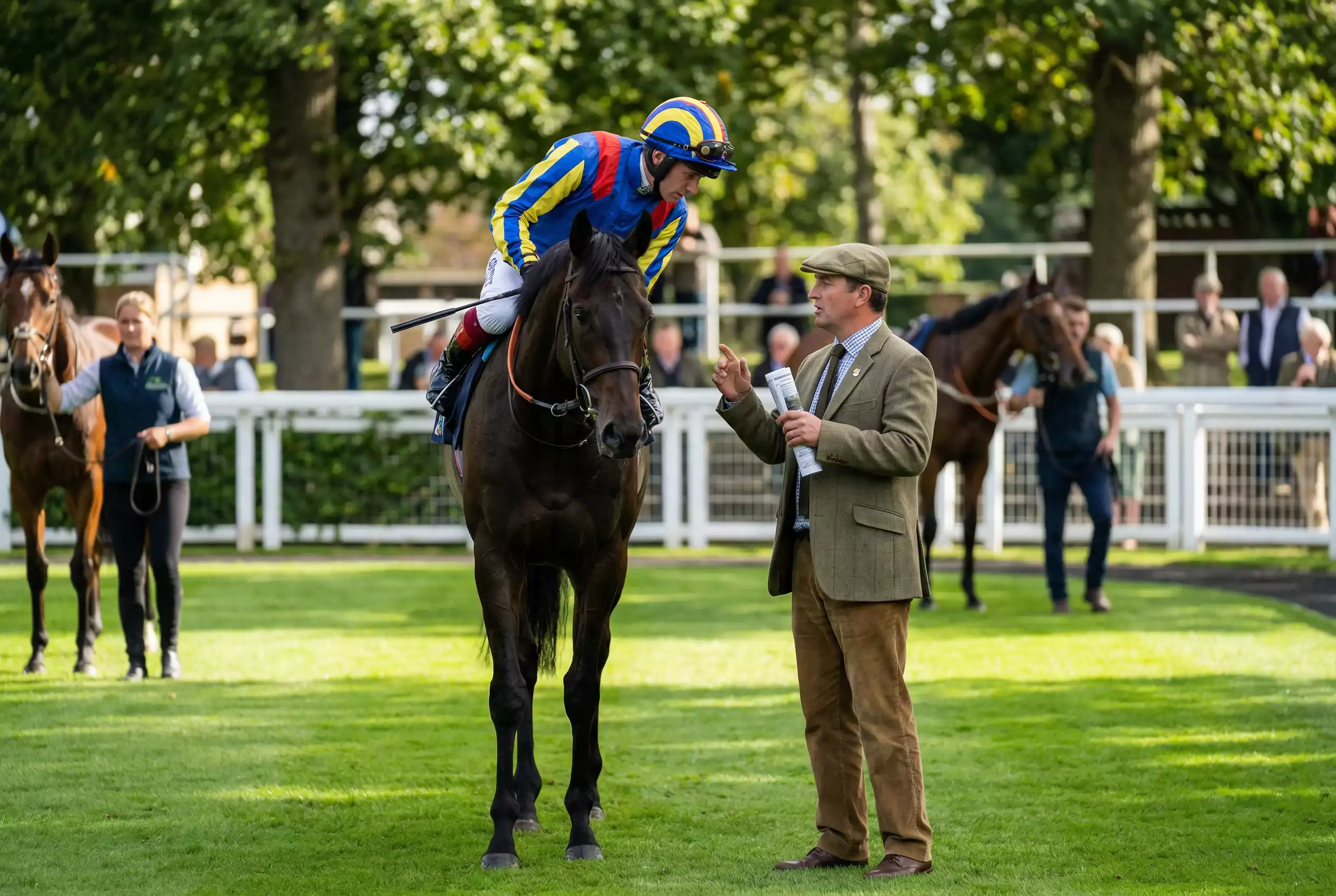 Jockey in colourful silks on horseback speaking with trainer at British racecourse paddock